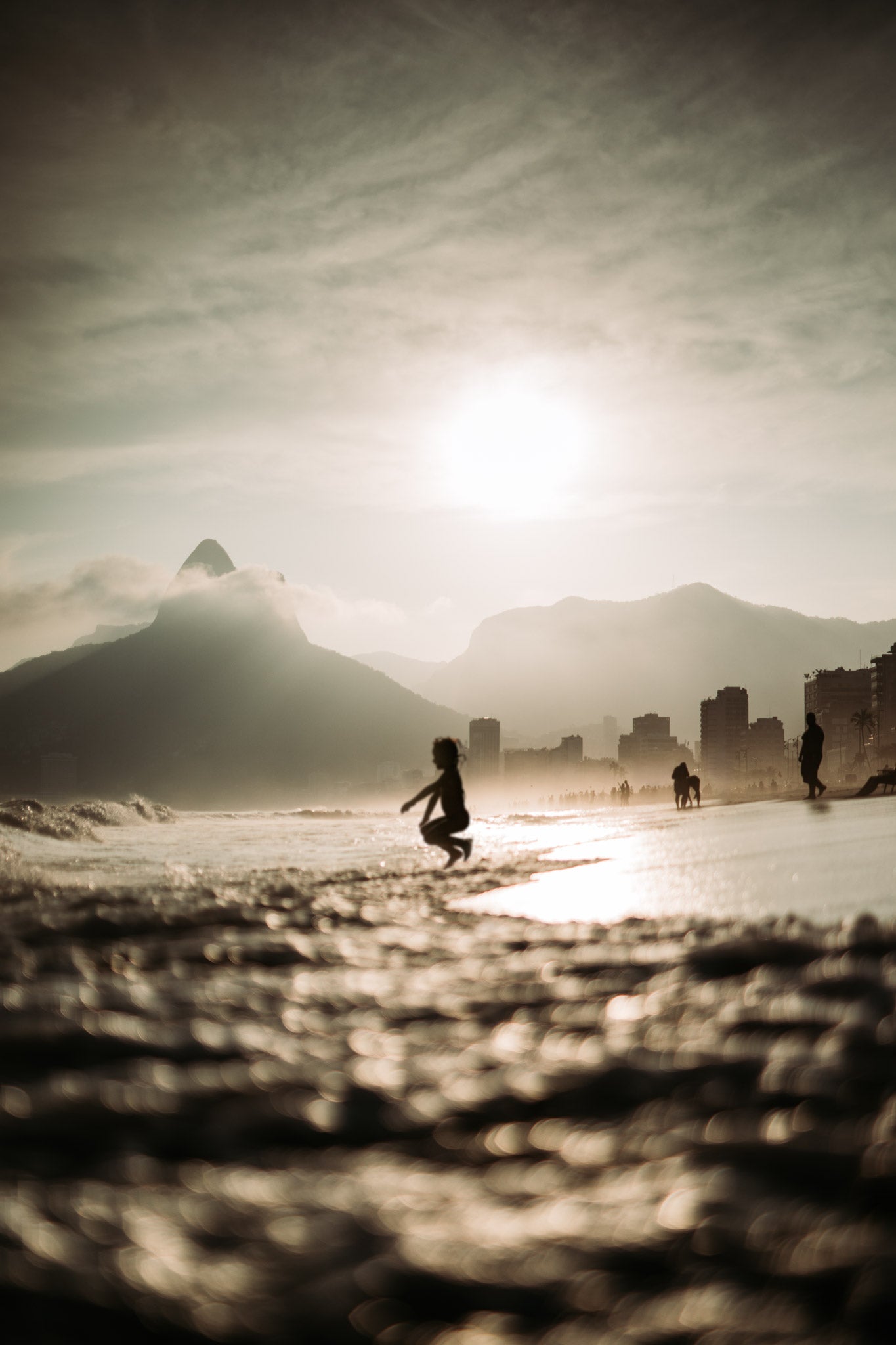 Ipanema Spring by Will Salkeld, silhouetted child jumping along the Ipanema shoreline at golden hour with Rio mountains behind, colour photography.