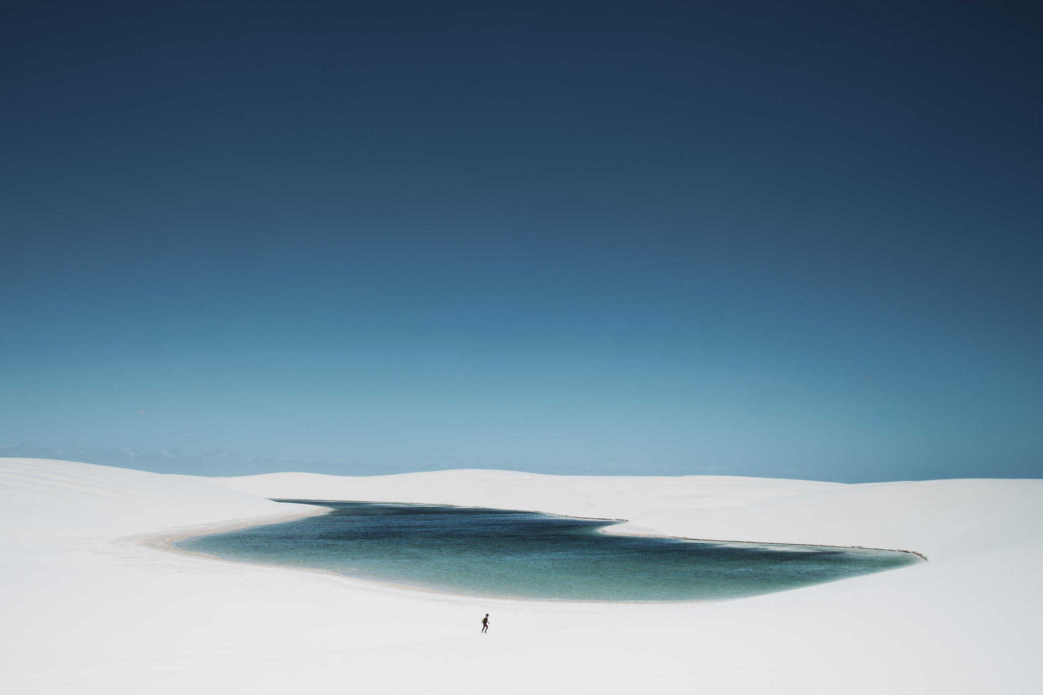 White Oasis by Will Salkeld, a small figure walking across white sand dunes toward a single turquoise lagoon at Lençóis Maranhenses, colour photography.
