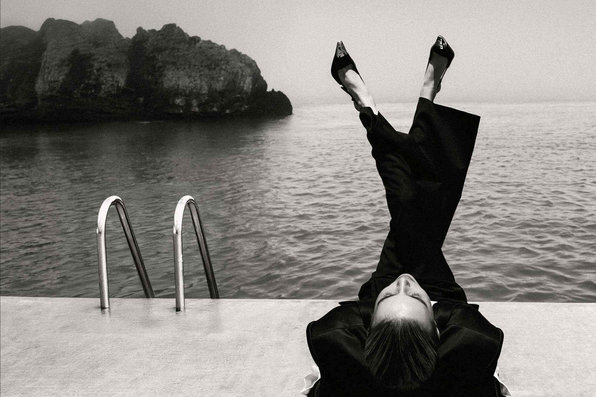 YSL by Raex Murillo, black and white photograph of a woman in black suit with legs raised vertically above a sea-edge pool platform