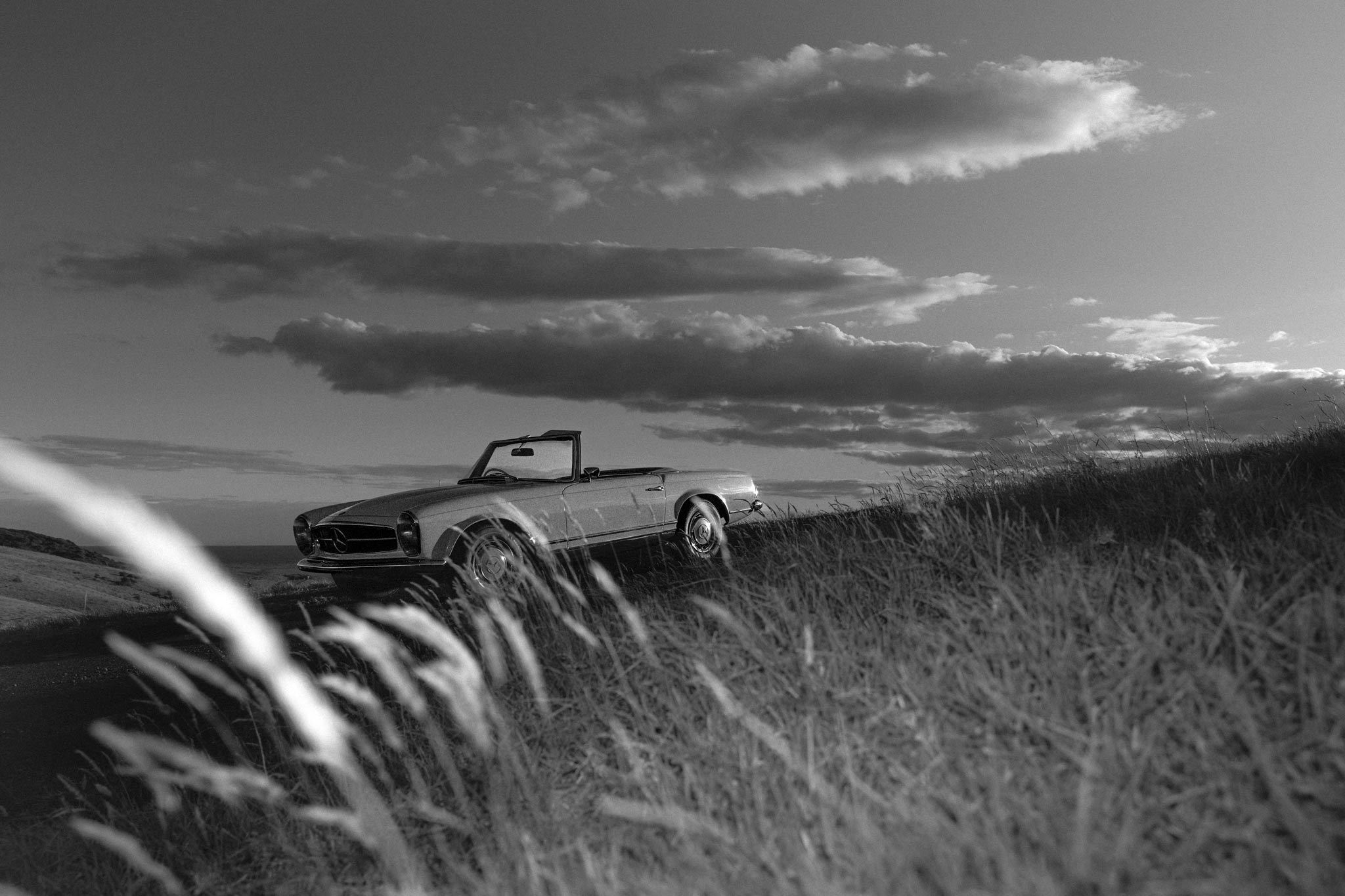 Cross Roads II by Raex Murillo, black and white photograph of a vintage Mercedes convertible on a coastal hill under stacked cloud