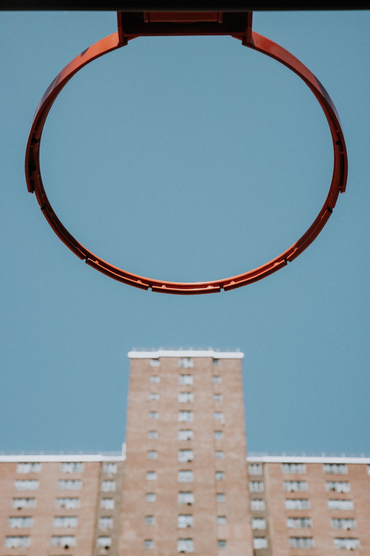 Perfect Shot by Will Salkeld, a red basketball hoop framed against blue sky above brick apartments at Rucker Park in Harlem, colour photography.