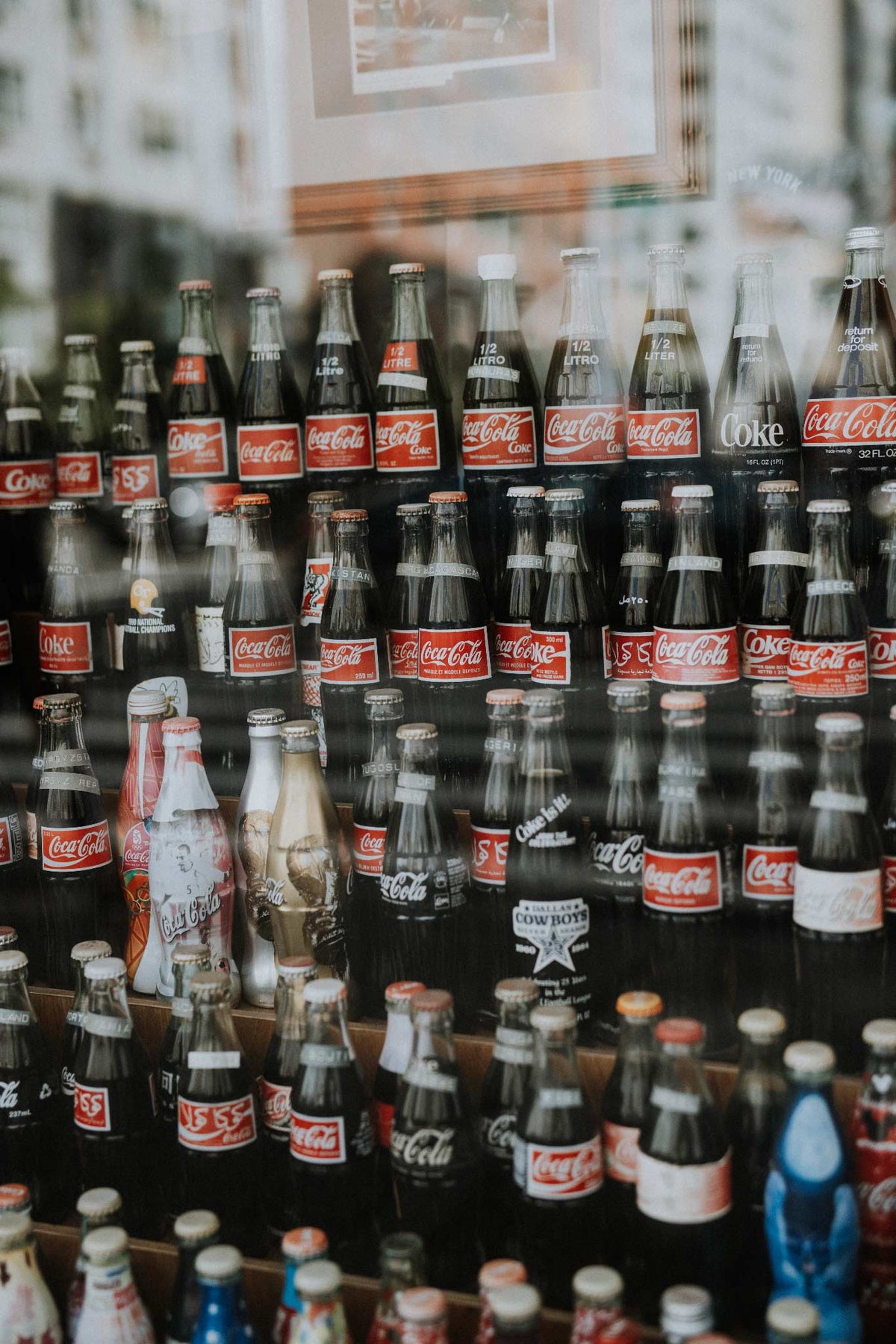 Coca Cola by Will Salkeld, rows of vintage Coca-Cola bottles displayed behind glass in a Manhattan soda shop, colour photography.