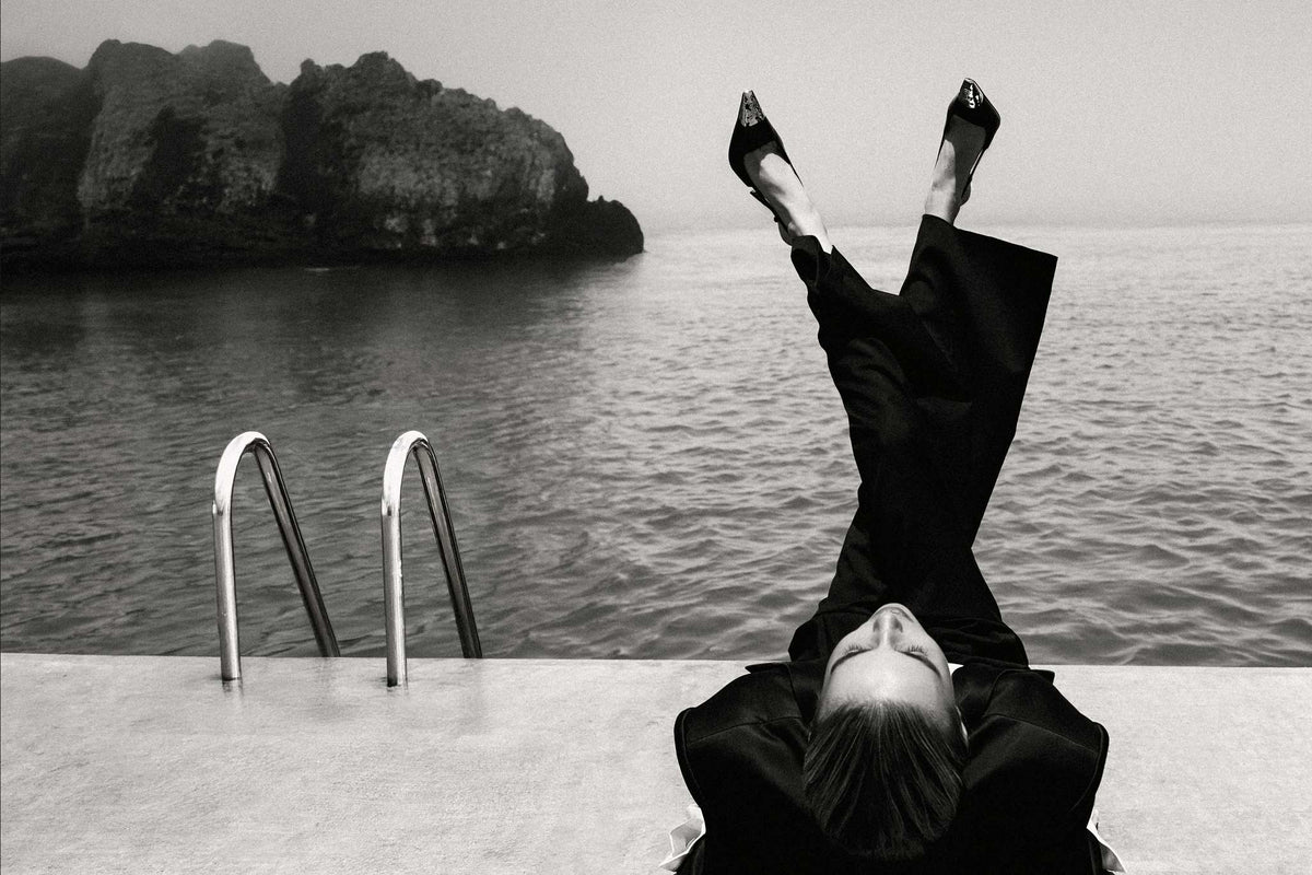 YSL by Raex Murillo, black and white photograph of a woman in black suit with legs raised vertically above a sea-edge pool platform