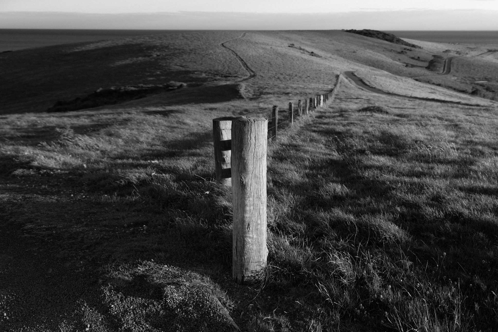 Cross Roads IV by Raex Murillo, black and white photograph of a single fence line running through a coastal hill toward the sea