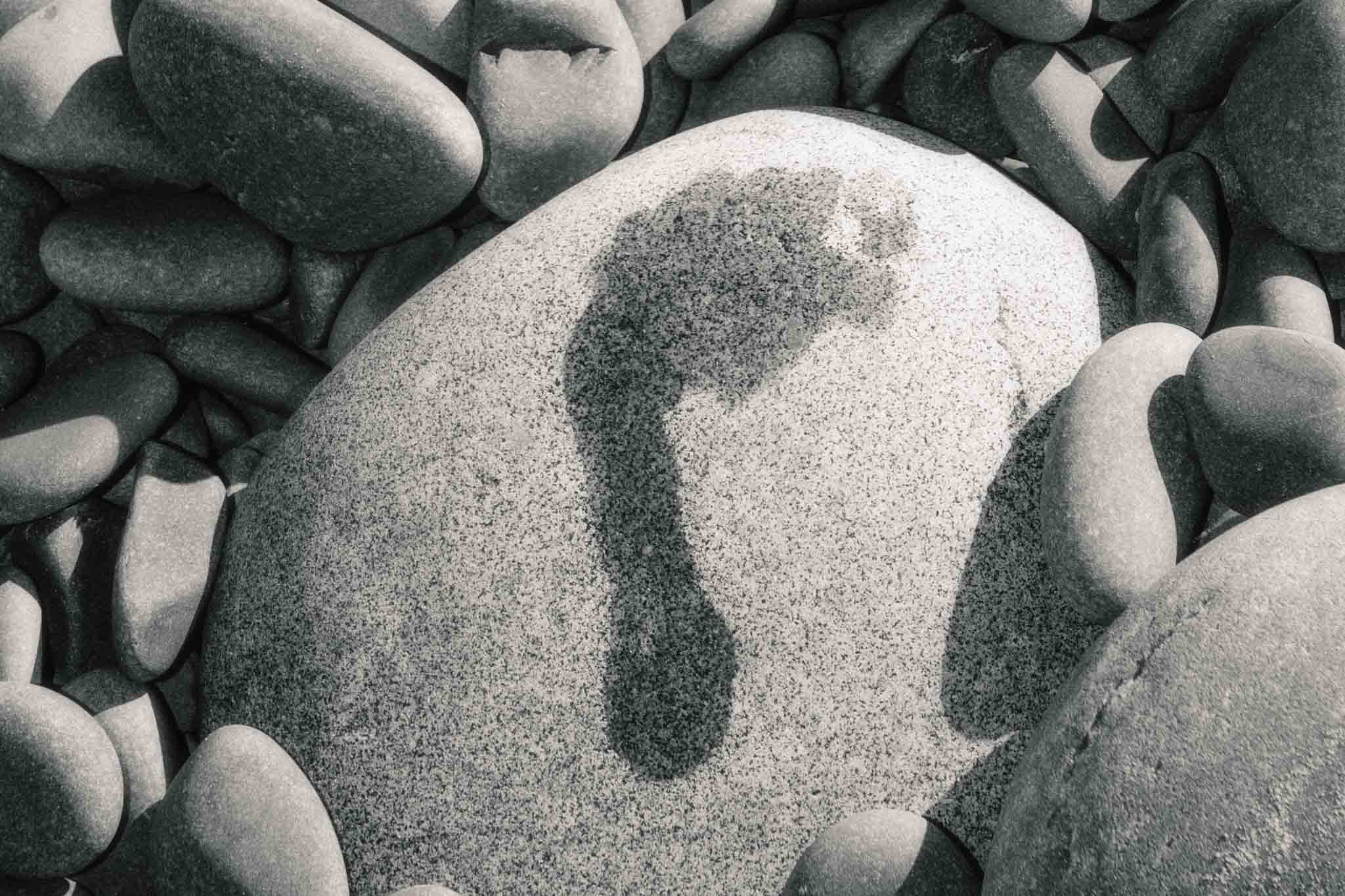 Stepping Stone by Raex Murillo, framed black and white landscape photograph of a single footprint pressed into a smooth tide-worn stone surrounded by sea-worn pebbles